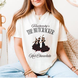 Könnte beinhalten: Ein cremefarbenes Kurzarm-T-Shirt mit schwarzem Text, der Tchaikovsky's THE NUTCRACKER und Hot Chocolate lautet. Unter dem Text befindet sich eine grafische Illustration von drei Tänzern in schwarzen Kleidern mit roten Akzenten beim Tanzen.