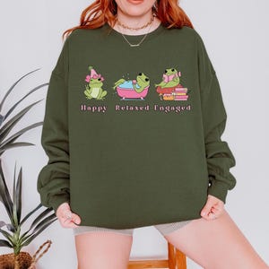 May include: Dark green sweatshirt featuring three cartoon frogs in different poses. The frogs are labeled "Happy", "Relaxed", and "Engaged". The first frog wears a pink hat, the second is in a pink bathtub, and the third reads a book.