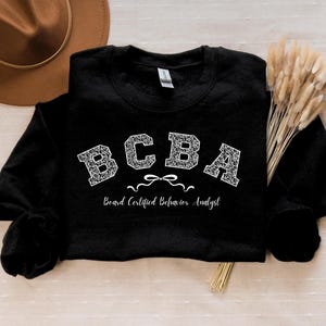 Può includere: Felpa nera con le lettere "BCBA" in un carattere decorativo. Sotto le lettere c'è il testo "Board Certified Behavior Analyst". Un cappello marrone e fiori secchi sullo sfondo.