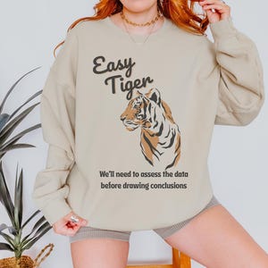 May include: Beige sweatshirt featuring the text "Easy Tiger" and a tiger illustration in brown and black. The text "We'll need to assess the data before drawing conclusions" is below the tiger.