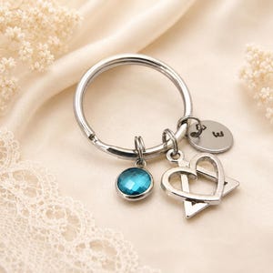 May include: A silver-toned metal keychain featuring three charms: a round faceted blue crystal, a silver heart shape layered over a triangle, and a small circular silver disc engraved with the letter E and a tiny heart symbol.