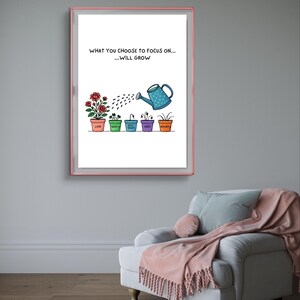 May include: A framed print with a pink border displays the text "WHAT YOU CHOOSE TO FOCUS ON... ...WILL GROW." Below, a watering can waters plants labeled "Love," "Worry," "Guilt," and "Anxiety." The print hangs above a white armchair.