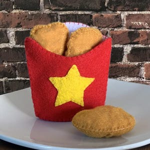 May include: A felt set of chicken nuggets on a white plate. The set includes four nuggets and a red holder with a yellow star. The text "CHICKEN NUGGETS" is displayed on the right.