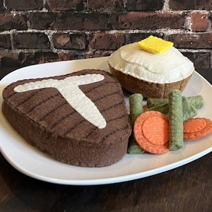 May include: A felt T-bone dinner set on a white plate. The set includes a brown T-bone steak with a white bone, a baked potato with a yellow butter pat, green beans, and carrot slices. The text reads "100% Handmade From Felt" and "T-BONE DINNER".