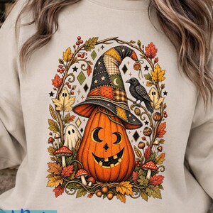 May include: A beige crewneck sweatshirt featuring a central graphic of an orange jack-o'-lantern wearing a patchwork witch hat. The design is surrounded by autumn leaves, small ghosts, mushrooms, and a black crow, all in a detailed illustration.
