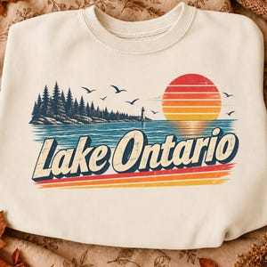 May include: A cream-colored crewneck sweatshirt featuring a graphic print of a landscape with a forest, a lighthouse, and a sunset over water. The text Lake Ontario is printed in a bold, dark blue font with a retro-style orange and red striped design.