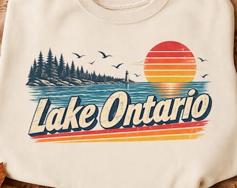 May include: A cream-colored crewneck sweatshirt featuring a graphic print of a landscape with a forest, a lighthouse, and a sunset over water. The text Lake Ontario is printed in a bold, dark blue font with a retro-style orange and red striped design.