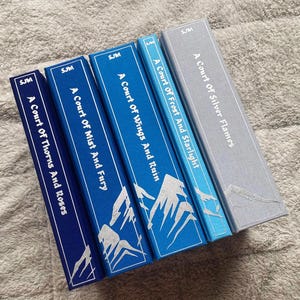 May include: Four books with the titles "A Court of Thorns and Roses", "A Court of Mist and Fury", "A Court of Wings and Ruin", and "A Court of Frost and Starlight" and "A Court of Silver Flames". The books have blue and grey covers with white text and mountain illustrations.