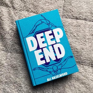 May include: A hardcover book titled "DEEP END" by Ali Hazelwood. The book cover is a vibrant turquoise with white lettering and line art illustrations of swimmers. The book is lying on a soft, gray textured surface.