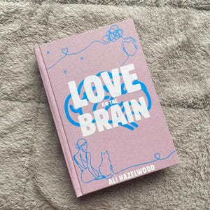 Puede incluir: Un libro de tapa dura rosa con el título "LOVE on the BRAIN" en texto blanco y un gráfico de cerebro azul. La portada presenta dibujos lineales azules de una persona, un gato y otras formas. El nombre del autor, Ali Hazelwood, está impreso en la parte inferior.