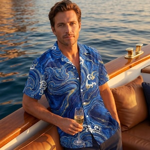 Op de afbeelding: Een overhemd met korte mouwen en knoopsluiting, voorzien van een levendig blauw en goud wervelend marmerpatroon met witte, golfachtige accenten. Het overhemd heeft een klassieke kraag en een knoopsluiting aan de voorzijde met witte knopen.