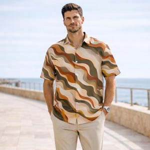 Puede incluir: Un hombre con una camisa de manga corta con un estampado ondulado en tonos marrón, beige y verde oliva, combinada con pantalones beige y zapatos de ante marrón. Camina por un paseo pavimentado junto al mar.