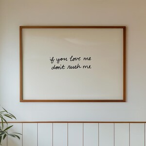 May include: A bathroom interior featuring a white bathtub, sink, and tiled walls. A framed print with the text "if you love me don't rush me" hangs above the tub. A small potted plant sits near the sink, adding a touch of greenery.