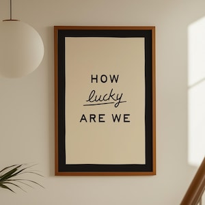 Puede incluir: Un cuadro enmarcado con las palabras "HOW lucky ARE WE" en escritura negra. La impresión tiene un fondo crema, un borde negro y un marco marrón. La obra de arte cuelga en una pared blanca cerca de una escalera.