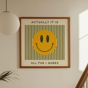 May include: A framed print featuring a yellow smiley face on a green striped background. The text "ACTUALLY IT IS" and "ALL FUN + GAMES" are displayed above and below the smiley face. The frame is a warm brown color.