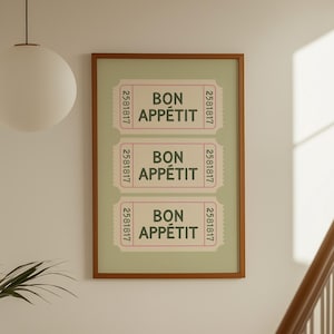 May include: A framed print featuring three pink and white ticket-style designs with the words "BON APPÉTIT" in the center. The print has a light green background and a brown frame. The print is hanging on a white wall.
