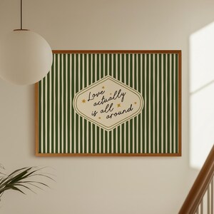 Puede incluir: Un cuadro enmarcado con un fondo a rayas verdes y blancas. La obra de arte presenta una forma de diamante color crema con el texto "Love actually is all around" en escritura cursiva. El marco es de color marrón cálido.