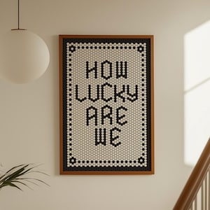 May include: Framed wall art with the text "HOW LUCKY ARE WE" in black. The artwork features a brown frame and a white and black tile design. A white globe light hangs from the ceiling, and a staircase is visible.