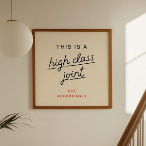 May include: A framed print with the text "THIS IS A high class joint ACT ACCORDINGLY" in black and red lettering. The print is in a brown frame and hangs on a white wall near a staircase and a hanging globe light.