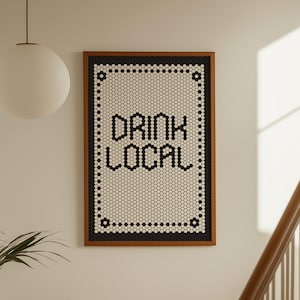 Peut inclure: Une œuvre d'art encadrée avec l'inscription "DRINK LOCAL" en lettres noires. L'œuvre présente un cadre marron et un fond blanc avec un motif de points noirs et blancs. L'art est accroché sur un mur blanc.