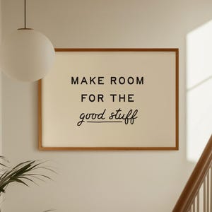 May include: A rectangular wall art print in a thin wooden frame featuring the black text "MAKE ROOM FOR THE good stuff" on a cream-colored background. The print is displayed on a plain wall, positioned above a staircase and near a hanging light.