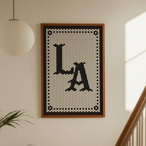 Puede incluir: Obra de arte enmarcada con las letras "LA" en negro sobre un fondo blanco de pequeñas baldosas hexagonales. El marco es de color marrón oscuro. La obra está colgada en una pared blanca, creando un contraste elegante.