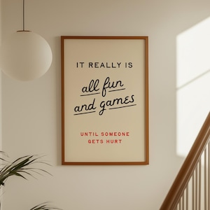 Pode incluir: Uma impressão emoldurada com o texto "IT REALLY IS all fun and games UNTIL SOMEONE GETS HURT" em preto e vermelho. A impressão está em uma moldura marrom e pendurada em uma parede branca perto de uma escada e uma luminária suspensa.