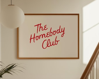 May include: A rectangular wall art print in a thin wooden frame featuring the red cursive text The Homebody Club centred on a white background. The artwork is displayed on a plain wall next to a staircase and a hanging globe pendant light.