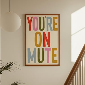 May include: A rectangular wall art print in a brown frame featuring the text YOU'RE ON MUTE in large, multi-colored sans-serif letters. The words are stacked in three rows with red, pink, olive green, light blue, and yellow typography.