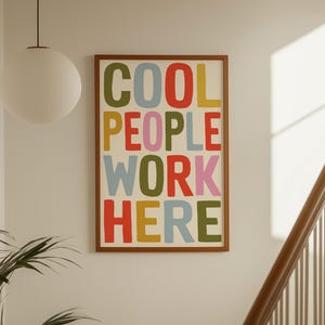 May include: A rectangular wall art print in a wooden frame featuring the text COOL PEOPLE WORK HERE in bold, multi-colored sans-serif letters. The words are stacked in four rows with alternating colors including olive green, red, yellow, and pink.