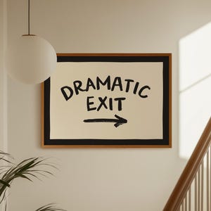 May include: A rectangular wall art piece in a wooden frame featuring a cream-coloured background with a thick black border. The text DRAMATIC EXIT is printed in bold black capital letters, accompanied by a black arrow pointing to the right.