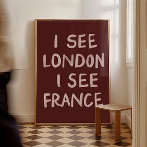 May include: A framed burgundy artwork with the text "I SEE LONDON I SEE FRANCE" in white. The artwork is in a room with a small wooden stool and a black and white checkered floor.