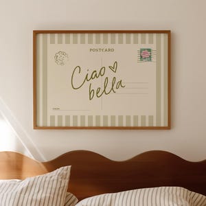 May include: A framed postcard print with the words "Ciao bella" in a handwritten style. The postcard has a beige background with vertical stripes and a postage stamp. The print hangs above a bed with striped bedding.