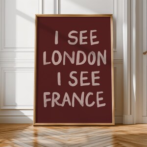 May include: A framed art print with the text "I SEE LONDON I SEE FRANCE" in a white, handwritten style on a deep red background. The frame is a light brown color. The artwork is displayed against a white wall and wooden floor.