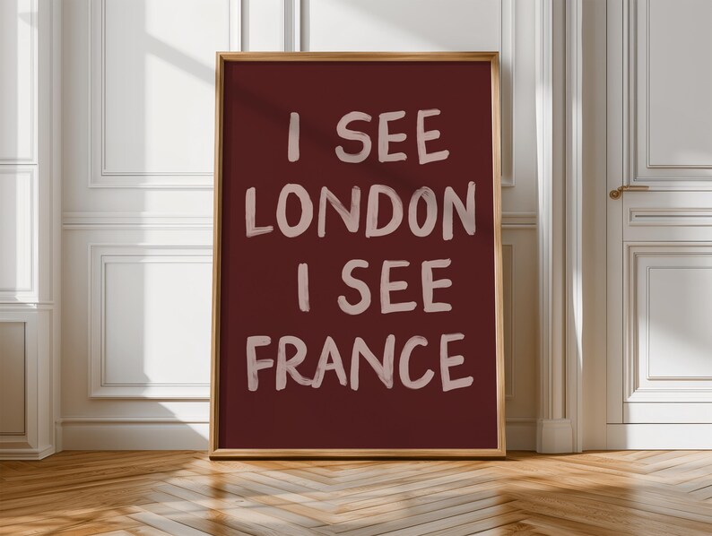 May include: A framed art print with the text "I SEE LONDON I SEE FRANCE" in a white, handwritten style on a deep red background. The frame is a light brown color. The artwork is displayed against a white wall and wooden floor.