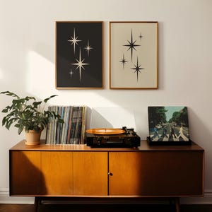 May include: Two framed prints with starburst designs hang above a mid-century modern cabinet. A record player with a vinyl record is on top of the cabinet, along with a stack of vinyl records and a potted plant.