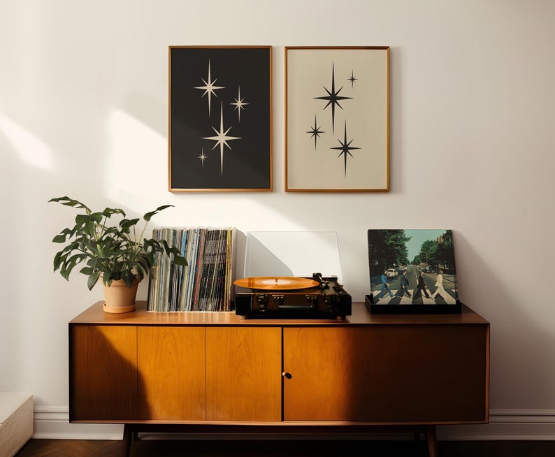 May include: Two framed prints with starburst designs hang above a mid-century modern cabinet. A record player with a vinyl record is on top of the cabinet, along with a stack of vinyl records and a potted plant.