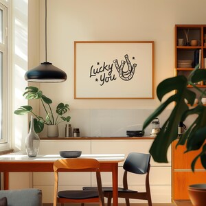 Może przedstawiać: Oprawiony wydruk z napisem "Lucky * You" i wzorem podkowy wisi nad stołem. W pokoju znajduje się czarna lampa wisząca, drewniane krzesła oraz szafka z książkami i dekoracjami. Rośliny dodają odrobinę zieleni.