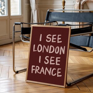 May include: A framed burgundy sign with the text "I SEE LONDON I SEE FRANCE" in white lettering. The sign is propped up on a wooden floor, with a black leather chair and a window in the background.