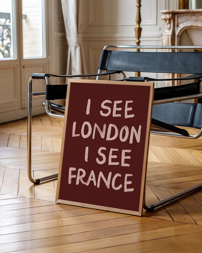 May include: A framed burgundy sign with the text "I SEE LONDON I SEE FRANCE" in white lettering. The sign is propped up on a wooden floor, with a black leather chair and a window in the background.