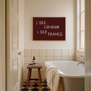 May include: A rectangular wall art piece with a dark maroon background and the white text I SEE LONDON I SEE FRANCE in a casual, hand-lettered font, displayed in a thin wooden frame above a white bathtub in a tiled bathroom setting.