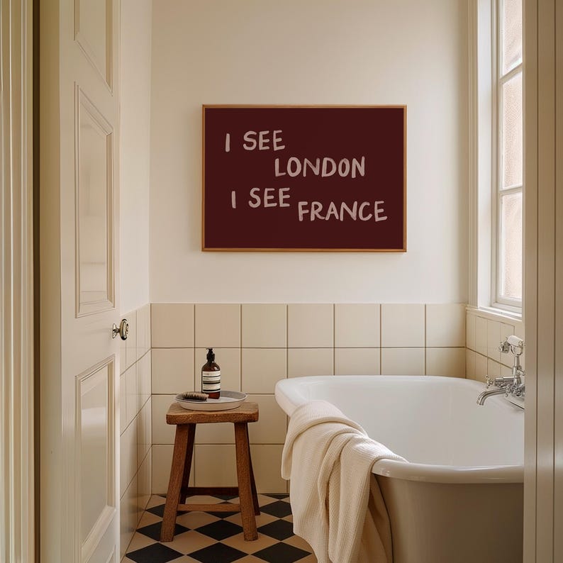 May include: A rectangular wall art piece with a dark maroon background and the white text I SEE LONDON I SEE FRANCE in a casual, hand-lettered font, displayed in a thin wooden frame above a white bathtub in a tiled bathroom setting.
