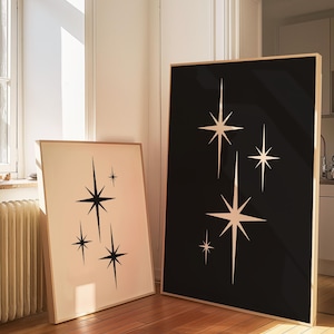 May include: Two framed art prints featuring starburst designs. One print has a black background with white stars, the other has a cream background with black stars. Both are in wooden frames and lean against a wall.