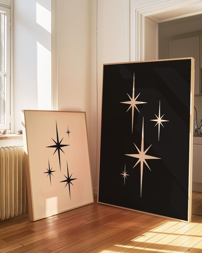 May include: Two framed art prints featuring starburst designs. One print has a black background with white stars, the other has a cream background with black stars. Both are in wooden frames and lean against a wall.