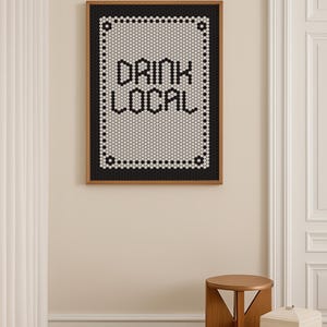 May include: A framed artwork with the words "DRINK LOCAL" in black letters. The text is set against a white background, surrounded by a black border, and framed in wood. The artwork is displayed on a beige wall.