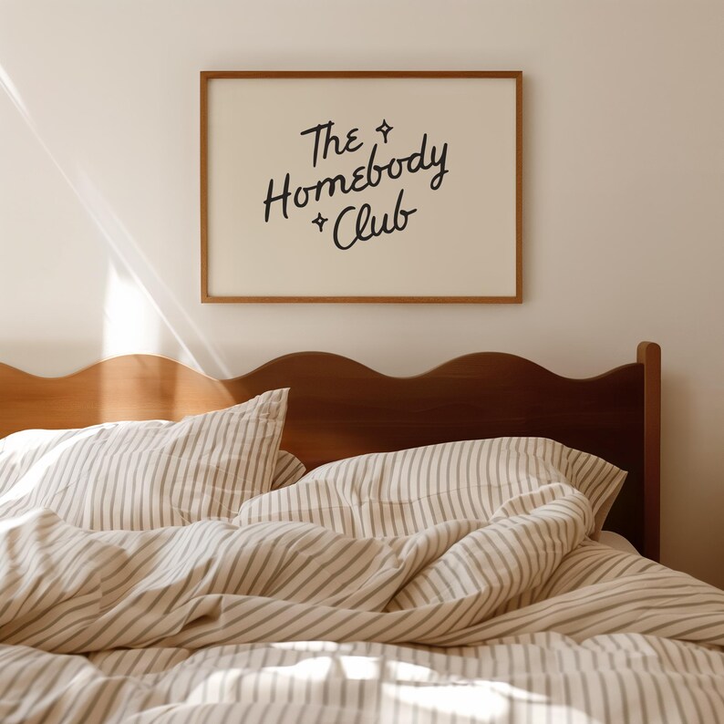May include: A framed print with the text "The Homebody Club" hangs above a bed with striped bedding. The print has a light background and a brown frame. The bedding is white with thin gray stripes. The headboard is made of dark wood.