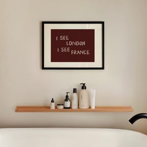 May include: A framed wall art print featuring a dark maroon background with the white text I SEE LONDON I SEE FRANCE. The print is displayed in a thin black frame with a cream-colored mat, positioned above a wooden shelf holding various bottles.