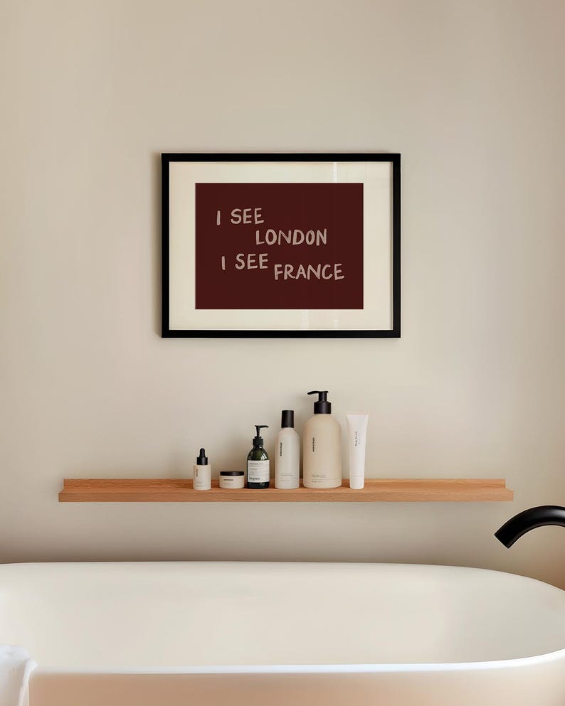 May include: A framed wall art print featuring a dark maroon background with the white text I SEE LONDON I SEE FRANCE. The print is displayed in a thin black frame with a cream-colored mat, positioned above a wooden shelf holding various bottles.