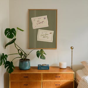 May include: A wooden dresser with a blue ceramic pot holding a large green plant. A framed board with a green and white striped pattern hangs on the wall, with two postcards that read "PS I Love you" and "Love You More!" on it.