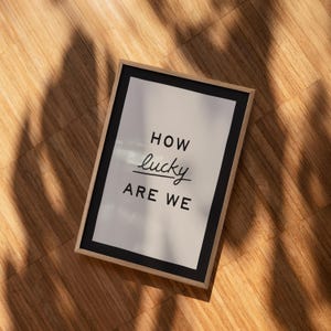 Puede incluir: Una impresi&oacute;n enmarcada con las palabras "HOW lucky ARE WE" en negro y fuente de escritura. La impresi&oacute;n tiene un borde negro y un fondo de color claro. El marco es de color madera clara, y la impresi&oacute;n est&aacute; sobre una superficie de madera.